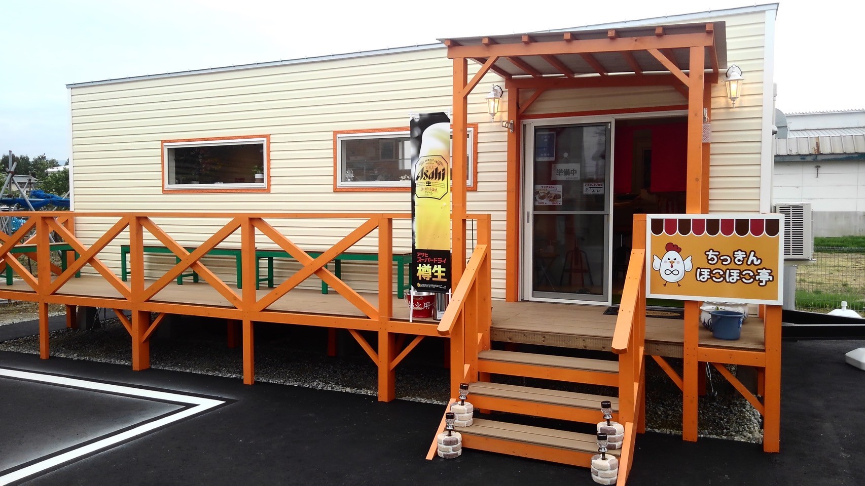 オレンジ色の外観と木のテラスが印象的なトレーラーハウス飲食店「ちっきんほこほこ亭」の外観。アサヒ樽生ののぼりと手書き看板が温かい雰囲気を演出